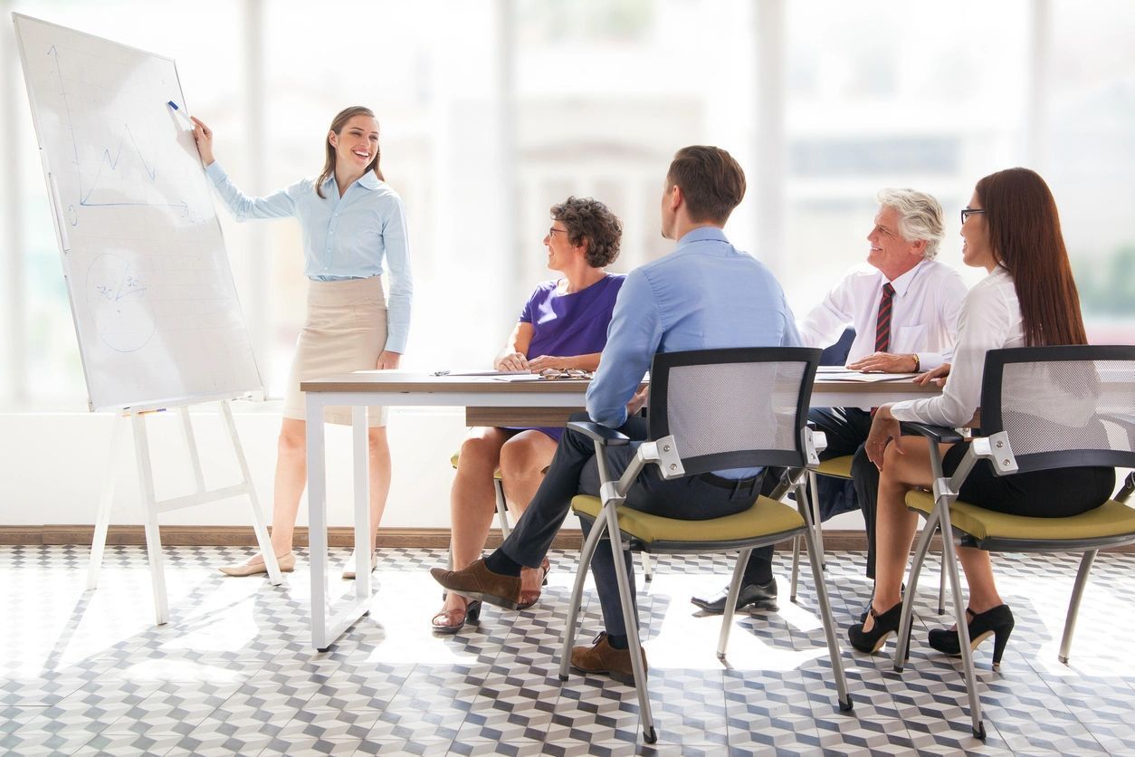 A woman standing at the front of a room with people sitting around.